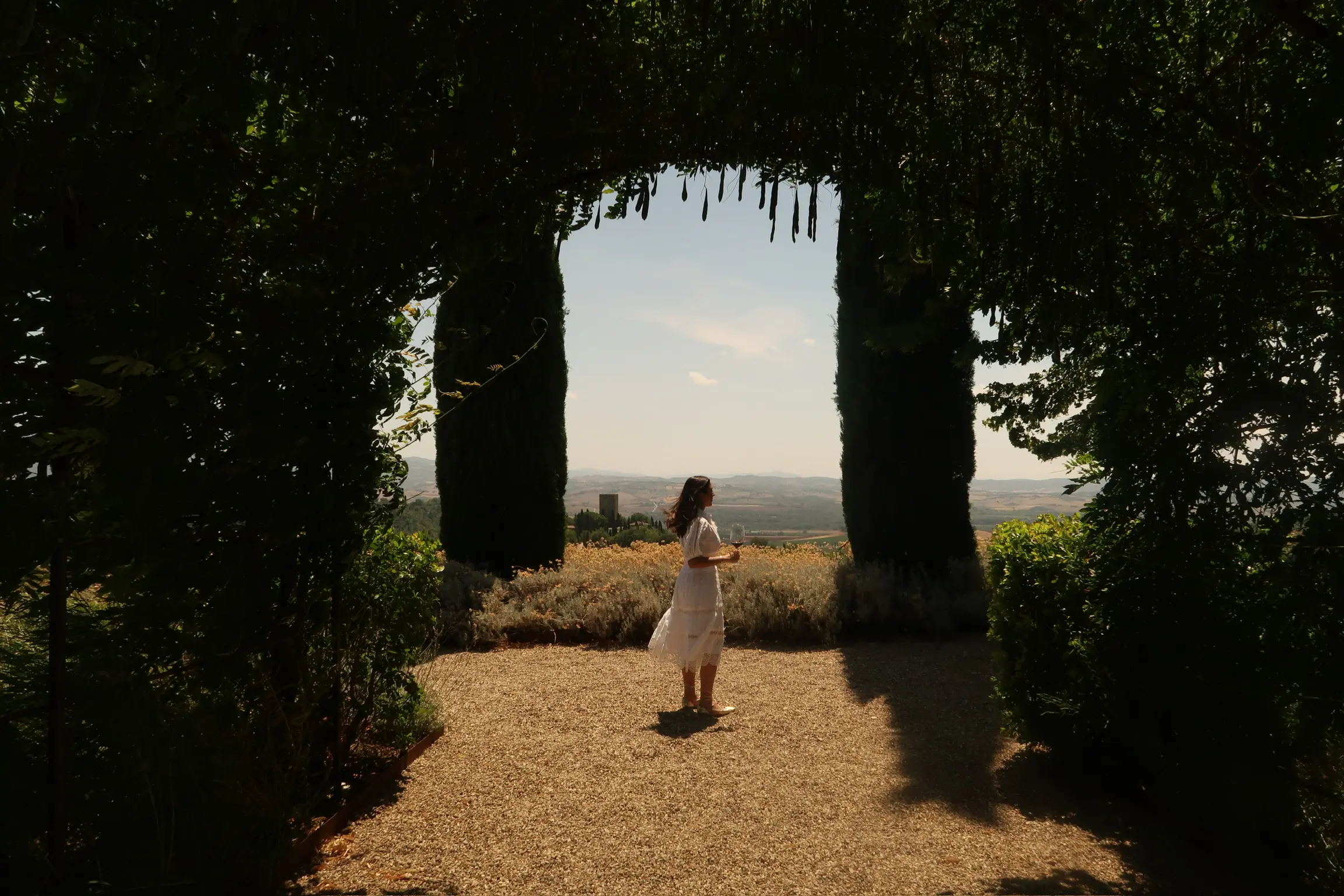 Viajera con vestido blanco y copa de vino bajo un arco de vegetación, ante la campiña toscana