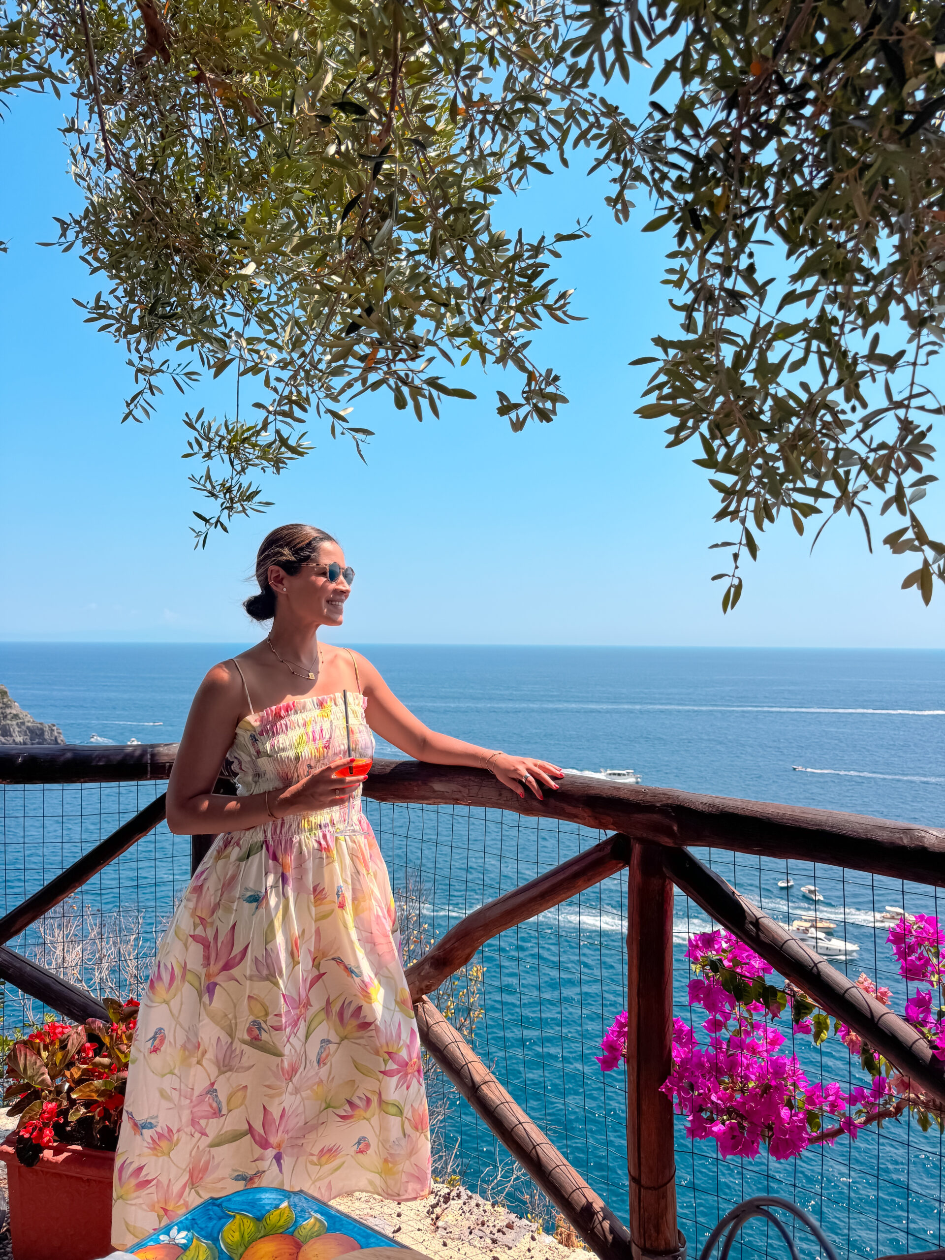 Keila Rodríguez República Dominicana Una mujer con un vestido floral y gafas de sol está de pie junto a una barandilla de madera, contemplando el océano.
