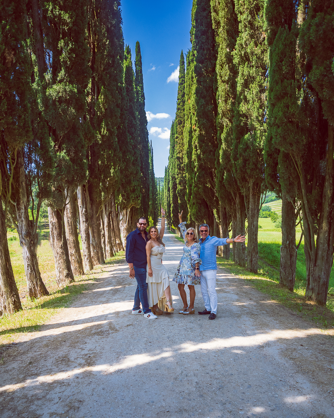 Un grupo de cuatro adultos sonrientes se encuentra en un camino soleado entre imponentes cipreses. Un grupo de cuatro adultos sonrientes se encuentra en un camino soleado entre imponentes cipreses.