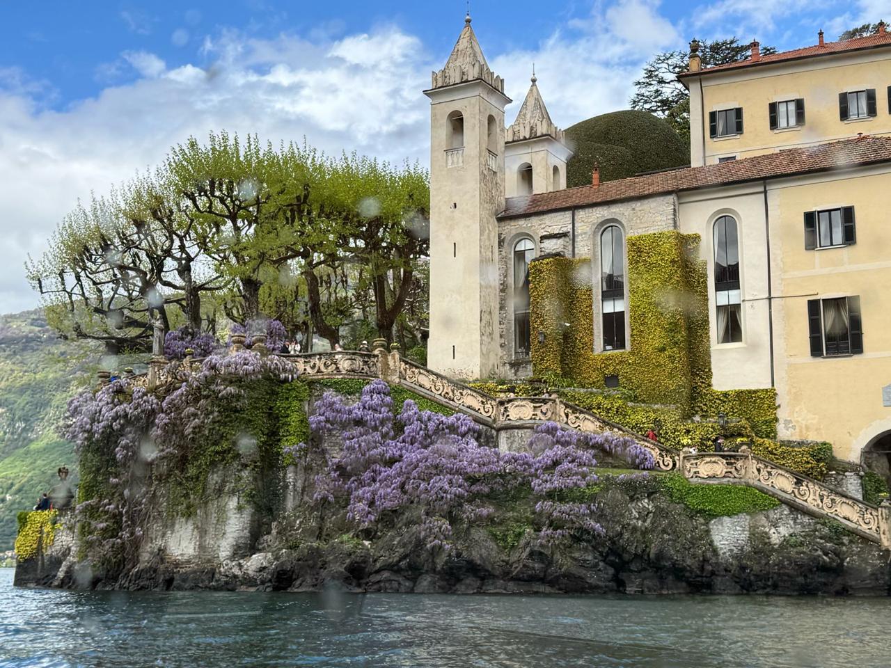 María Isabel y Ho Chi Vega México Elegante villa junto al lago con paredes cubiertas de hiedra, una alta torre y una ornamentada escalera adornada con exuberantes glicinas moradas.