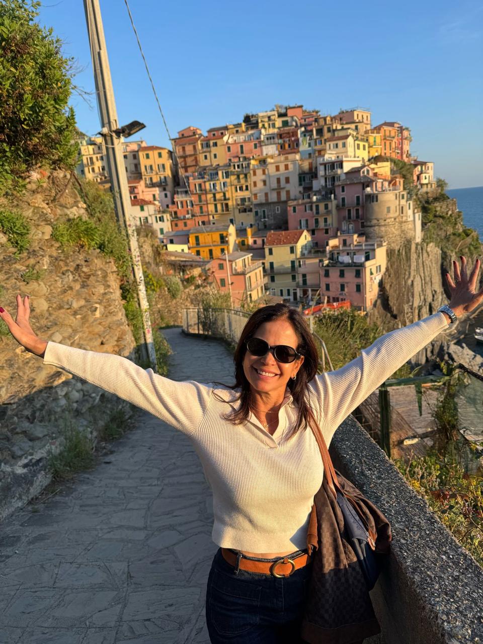 María Isabel y Ho Chi Vega México Una mujer con gafas de sol extiende alegremente los brazos, de pie en un camino con coloridos edificios en la ladera y un cielo azul brillante al fondo.