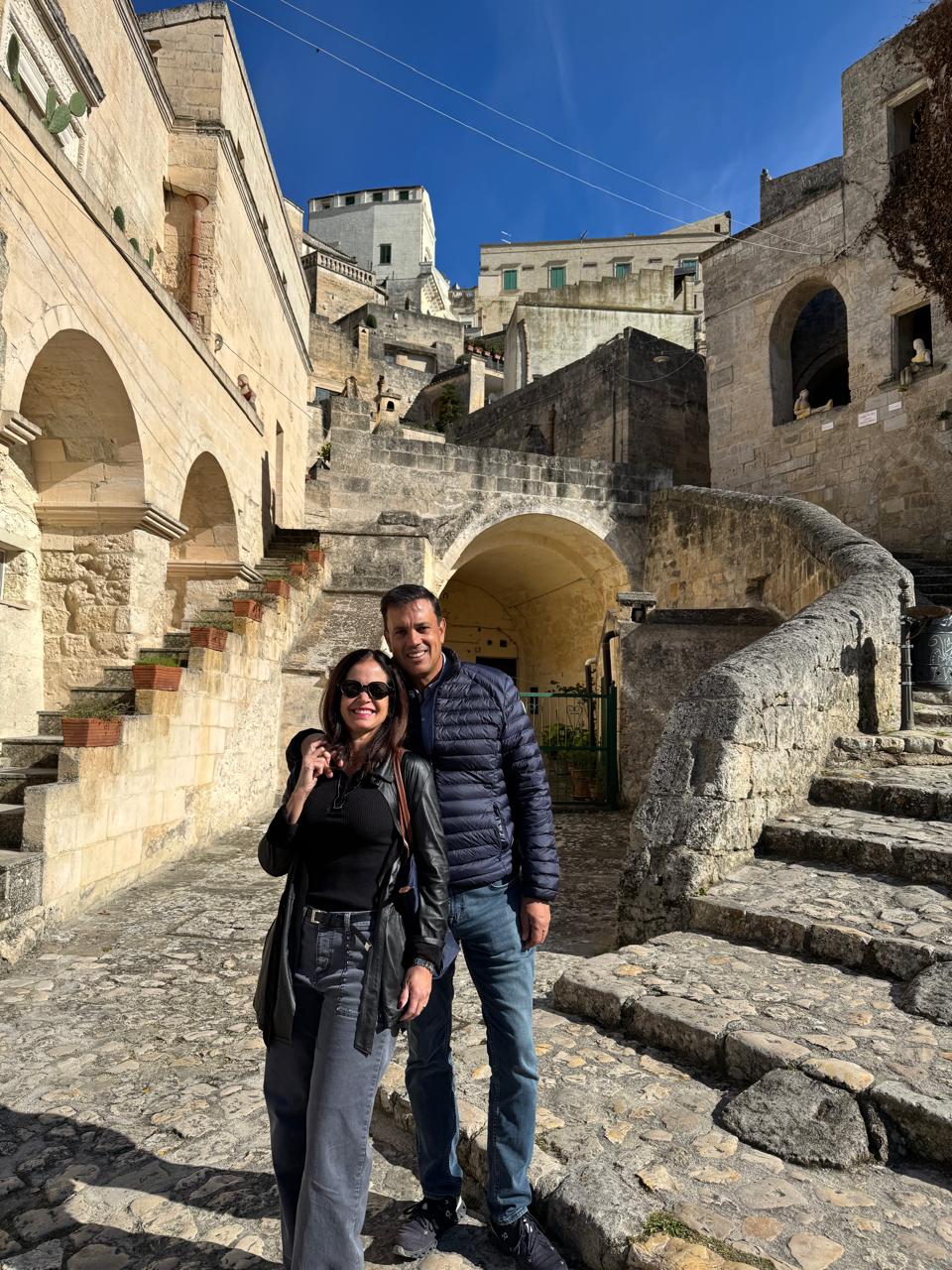 María Isabel y Ho Chi Vega México Una pareja sonriente posa en unos escalones de piedra frente a unos edificios históricos bajo un cielo azul claro, irradiando calidez y satisfacción.