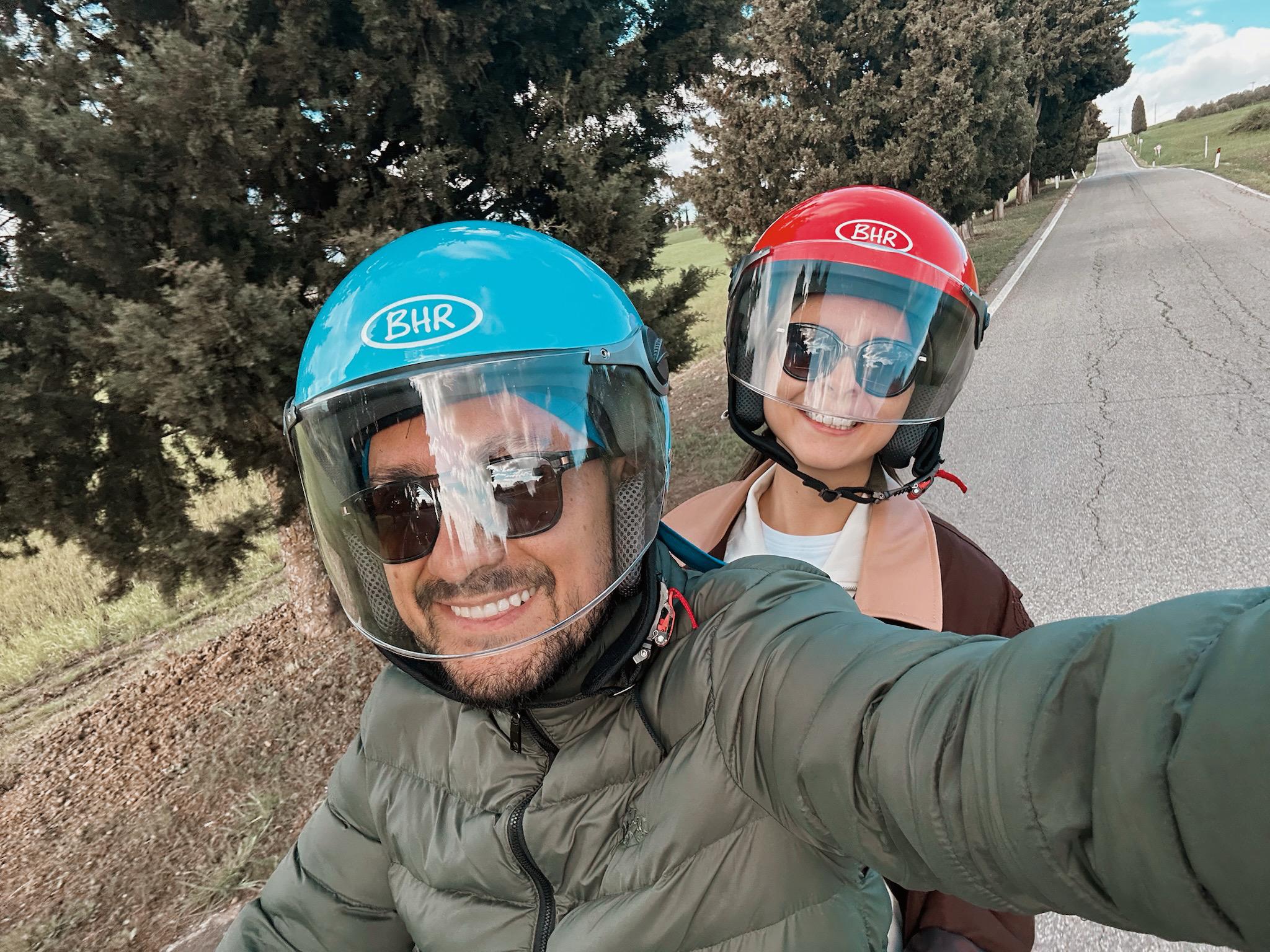 Lula & Oscar Cortés Ecuador Una pareja sonriente con cascos, uno azul y otro rojo, se hace un selfie en una carretera rural.
