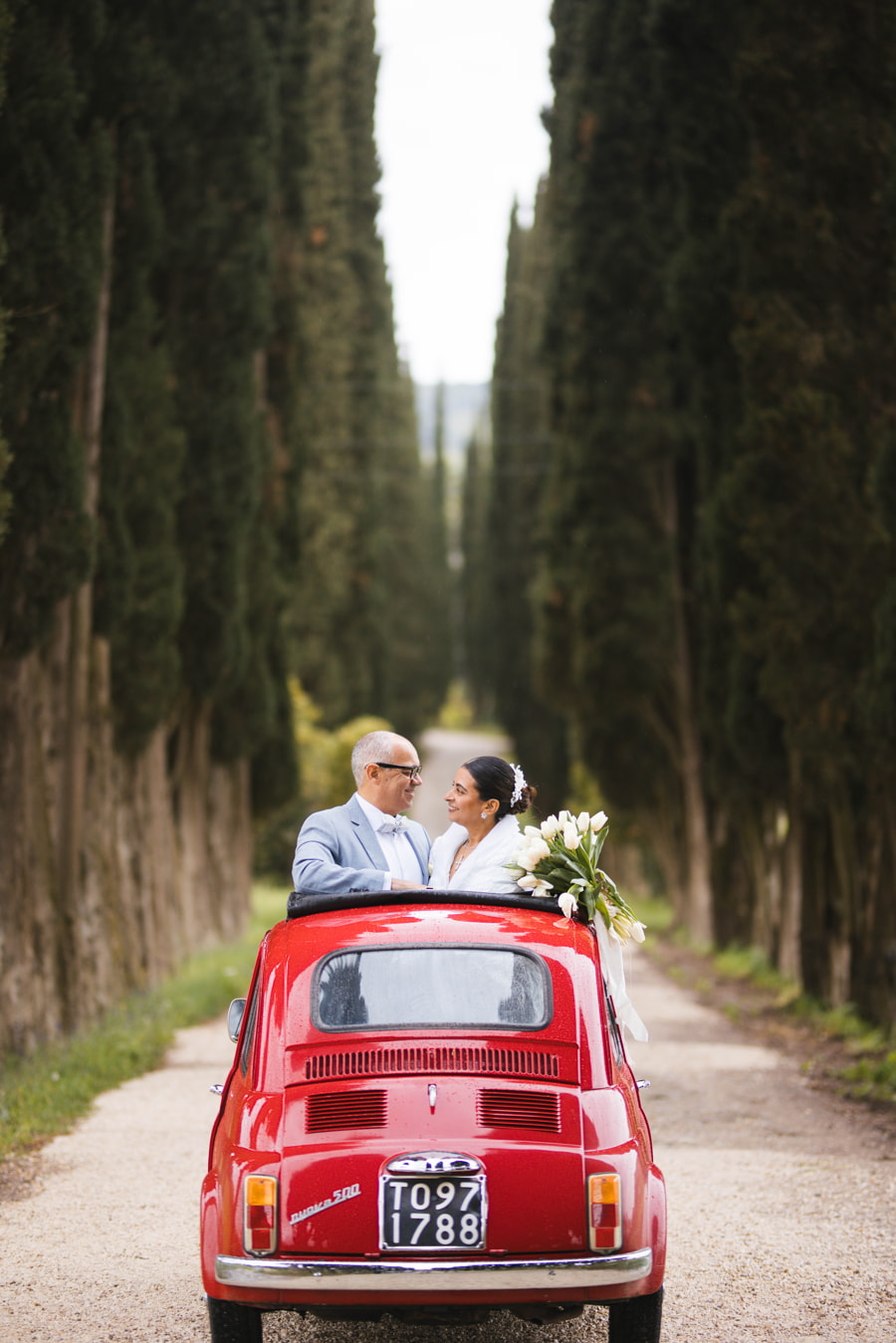 Una pareja se sonríe alegremente desde un coche rojo antiguo en una carretera bordeada de árboles.