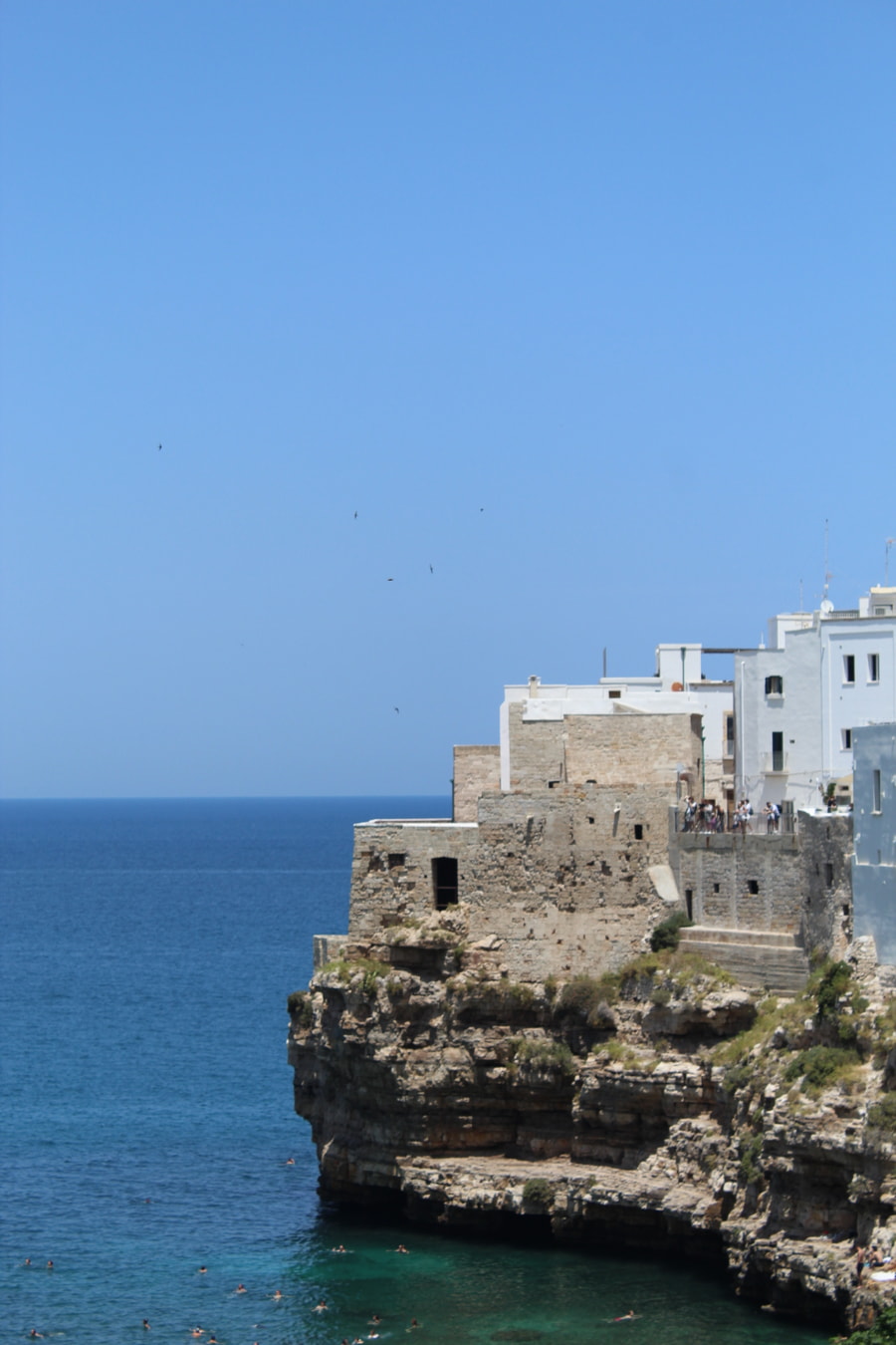 Vista desde un acantilado de una ciudad costera con edificios de piedra y encalados que se asoman a un mar azul profundo bajo un cielo despejado.