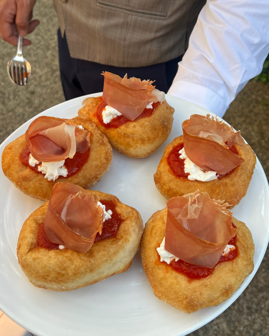 Un camarero presenta un plato con cinco pequeños discos de masa frita cubiertos con salsa de tomate, ricotta y una loncha de embutido.