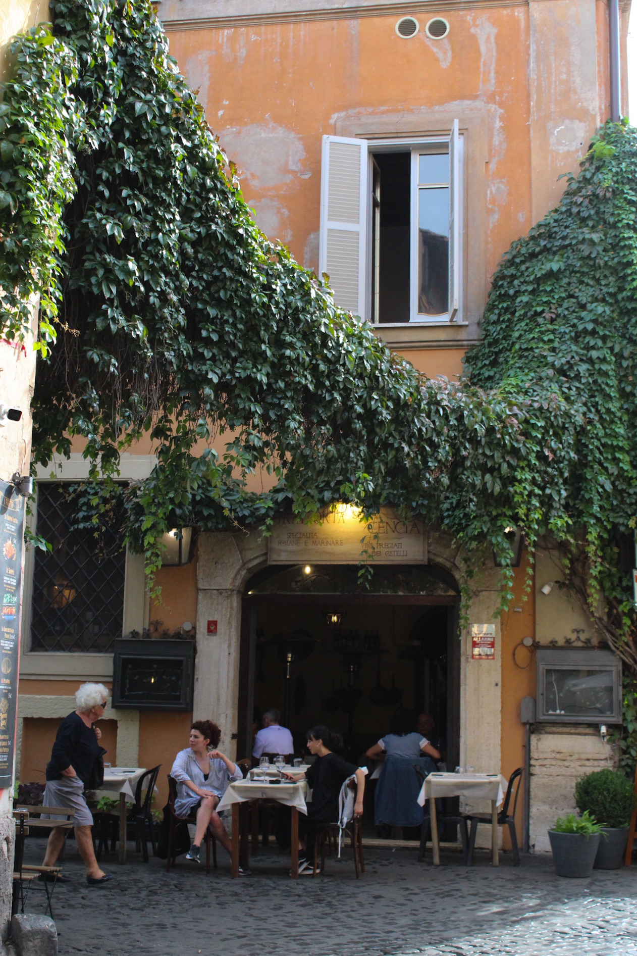 Trattoria tradicional en Roma Trattoria romana con fachada naranja cubierta de hiedra y comensales en mesas al aire libre sobre una calle empedrada