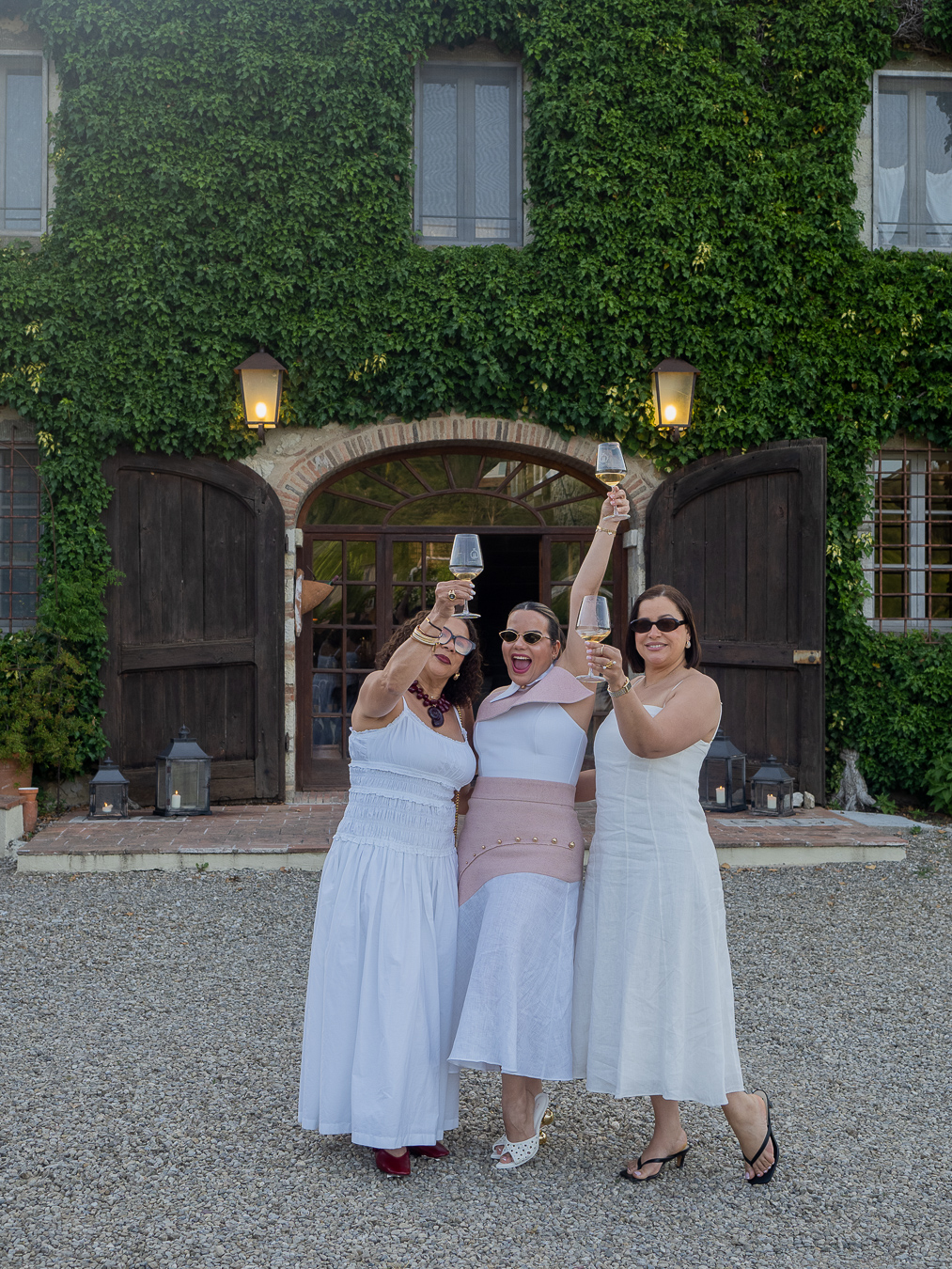 Tres amigas brindando con vino blanco frente a una villa toscana cubierta de hiedra