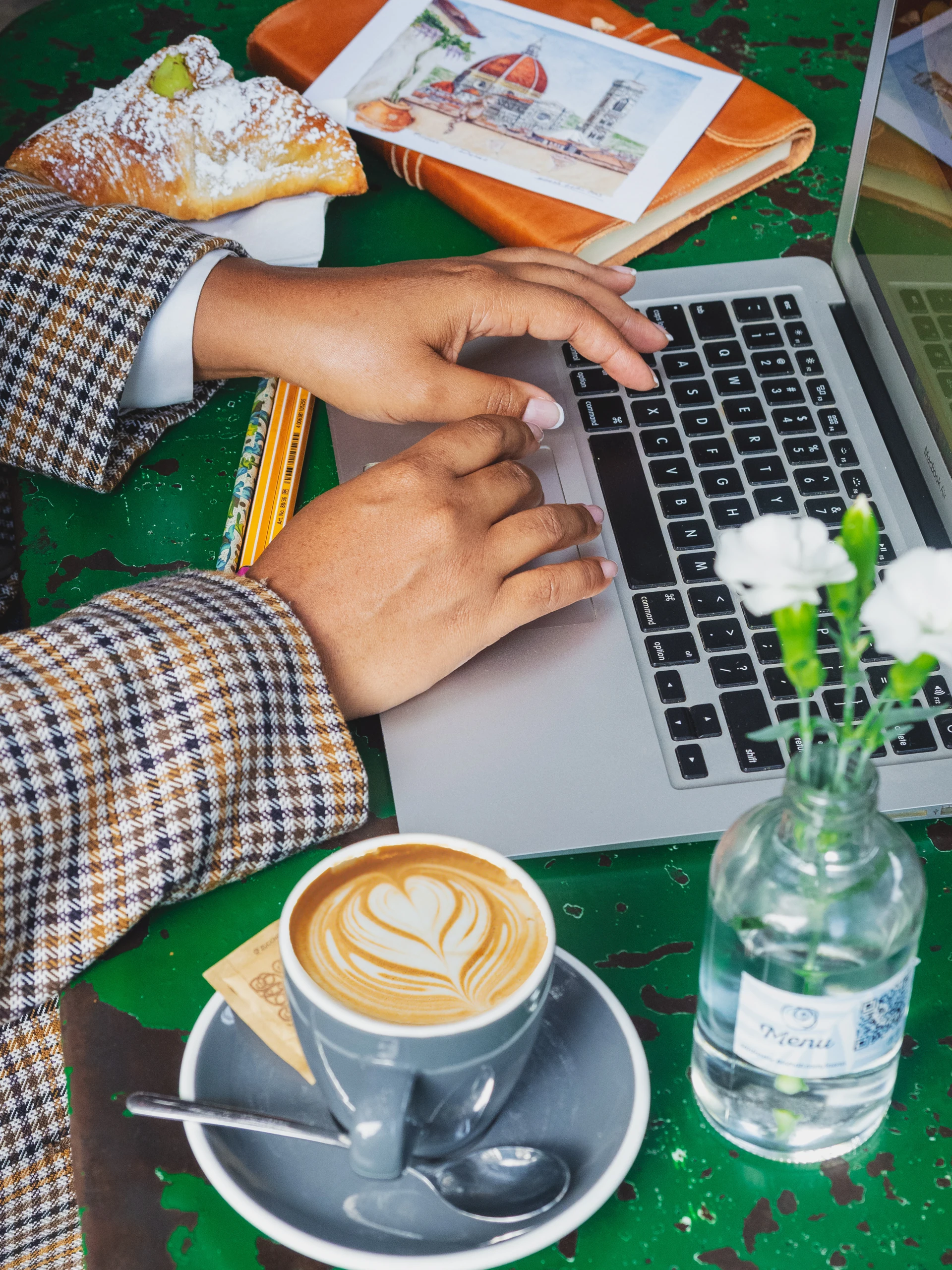 Diseñando un viaje personalizado a Italia Manos escribiendo en un portátil junto a un capuchino, un cornetto y una acuarela del Duomo de Florencia